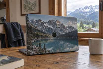 MacBook-Skin mit einem Bergsee-Foto als Motiv, auf einem Holztisch vor Bergpanorama. AI-generiertes Bild.